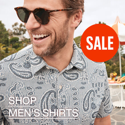 A close up image of a man wearing a light blue polo with a paisley print in dark blue. He is wearing sunglasses and the words Shop Men's Shirts read across the bottom left of the image. 